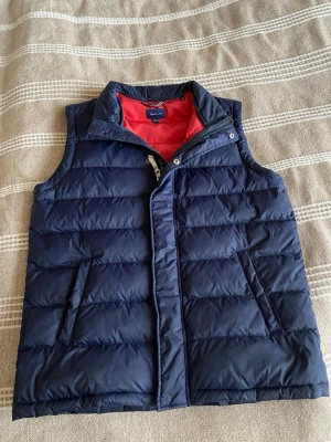 Mörkblå dunväst från GANT - Snygg mörkblå dunväst från GANT med röd insida och ståkrage. Västen har två fickor med dragkedja och stängs med både dragkedja och tryckknappar. Perfekt för lager på lager och ger en sportig look.