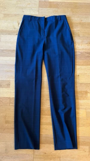 Mörkblå rutiga kostymbyxor - Snygga mörkblå kostymbyxor med diskret rutmönster. Byxorna har raka ben, pressveck och sidfickor fram samt passpoalfickor bak. Perfekta för en stilren look. Materialet känns slätt och klassiskt, troligen polyester eller ullblandning.