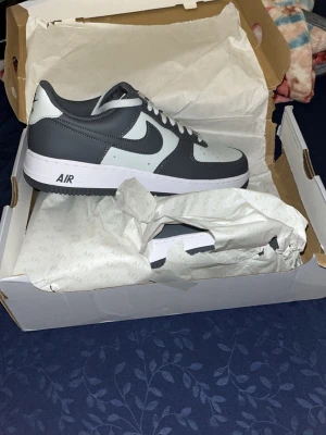 Nike Air Force 1 grå/vit sneakers - Snygga Nike Air Force 1 sneakers i grått och vitt. Klassisk låg modell med vit sula och mörkgrå detaljer. Ikonisk Swoosh-logga på sidan och AIR-tryck på sulan. Perfekta för dig som vill ha en stilren och tidlös sneaker i skinn.