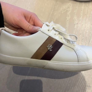 Vita sneakers från Ralph Lauren - Snygga vita sneakers från Ralph Lauren med bruna och mörkbruna ränder på sidan samt RL-logga i metall. Skorna har klassisk låg profil, vita snören och ovandel i slätt konstläder. Perfekta för en clean och stilren look. Använda två gånger men är för stora för mig. Skulle gissa på att de passar den som har 38/38,5