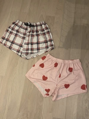 Två par pyjamasshorts med mönster - Säljer två par pyjamasshorts, ett par i rosa med röda hjärtan och ett par i vitt med rött och svart rutmönster. Båda har elastisk midja och är perfekta för mysiga kvällar hemma.