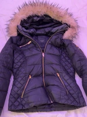 Svart pufferjacka med fuskpäls - Snygg svart pufferjacka med guldfärgade dragkedjor och quiltade detaljer. Jackan har en stor huva med beige fuskpäls och flera praktiska fickor. Perfekt för kalla vinterdagar och ger en trendig look.