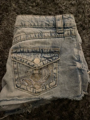 Ljusblå jeansshorts med paljettdetaljer - Säljer ett par ljusblå jeansshorts med rå kant och  paljettdetaljer på bakfickorna. 