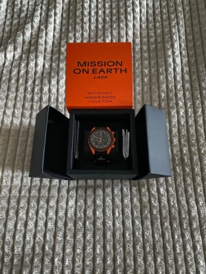 Omega x Swatch Mission on Earth Lava - Säljer en Omega x Swatch Mission on Earth Lava från Bioceramic MoonSwatch Collection. Klockan har en svart urtavla med vita och orange detaljer, samt en orange boett och svart rem med Omega-logga. Materialet är biokeramik och remmen är syntet.