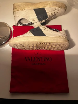  Äkta Valentino Garavani open vita och mörk blåa sneakers - Snygga vita sneakers från Valentino Garavani med bred svart läderdetalj över sidan. Klassisk låg modell med rund tå och vita snören. Yttermaterial i läder och gummisula. Perfekta för dig som vill ha en stilren och exklusiv look. Nypris är ca 7000kr men jag har endast använt dom i en sommar . Priset är inte hugget i sten så hör av er vid funderingar. MVH Eric 