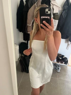 Vit kort klänning - Säljer en söt axelbandslös kort klänning med figurnära passform och ett snyggt rosett knyte i ryggen. Aldrig använd och prislapp sitter kvar. Skriv för fler bilder💕