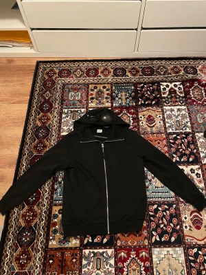 Svart hoodie från C.P. Company - Säljer en svart hoodie från C.P. Company med dragkedja framtill och klassisk huva med inbyggda glasögon. Hoodien har en stilren look och är perfekt för dig som gillar streetwear. Materialet känns mjukt och bekvämt, och passformen är avslappnad.