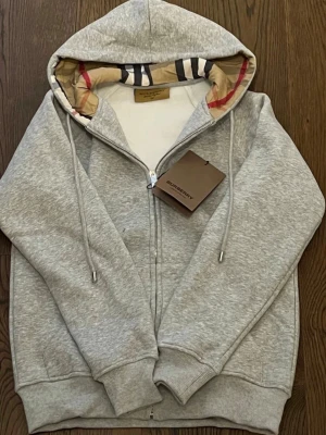 Grå zip-hoodie från Burberry - Snygg grå hoodie med dragkedja från Burberry. Klassisk huva med det ikoniska rutmönstret på insidan, dragsnören och ribbade muddar. Perfekt för en chill och stilren look. Materialet är mjukt och bekvämt, passar till det mesta.