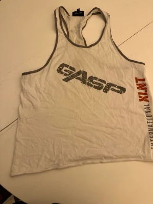 Vit linnetopp från GASP - Säljer en vit linnetopp från GASP med grå kantband och tryck i svart och rött framtill. Toppen har ärmlös design och sportig look, perfekt för träning eller gym. Materialet är mjukt och lätt bomull som andas.