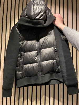 Svart pufferjacka från BVALDI - Snygg svart pufferjacka från Bvaldi,  med stickade ärmar och sidor, quiltad front och stor huva. Jackan har två dragkedjeförsedda fickor och en metallpatch med Bvaldi-logga på ärmen. Perfekt nu inför våren! Pris går att diskutera. 