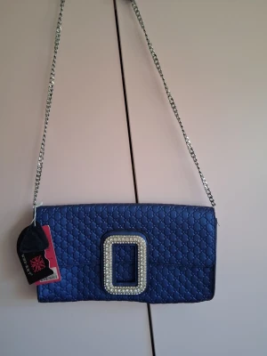 Blå quiltad kuvertväska med kedja - Snygg blå kuvertväska från med quiltat mönster och en stor, glittrig spännesdetalj framtill. Väskan har en silverfärgad kedjerem och elegant design som sticker ut. Perfekt för dig som vill ha något extra till din outfit.