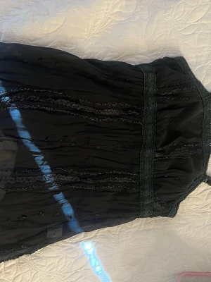 Svart tröja med spetsdetalj - Säljer en svart tröja  med tunna axelband och fina spetsdetaljer i midjan. Tyget är lätt och har subtila dekorativa sömmar som ger en unik look. Perfekt för dig som gillar en stilren men ändå detaljrik outfit.