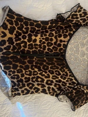 Leopardmönstrad mesh-top - Snygg top i transparent mesh med leopardmönster. Svart spetskant runt urringningen och korta ärmar med volangdetalj. Perfekt för dig som vill sticka ut med ett djurmönstrat plagg. Ingen synlig logga eller märke.