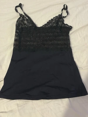 Svart spetsnattlinne med justerbara band - Snyggt svart nattlinne med spetsdetaljer upptill och släta, mjuka tyg nedtill. Justerbara axelband för perfekt passform och v-ringad halsringning. Perfekt för en mysig kväll hemma.