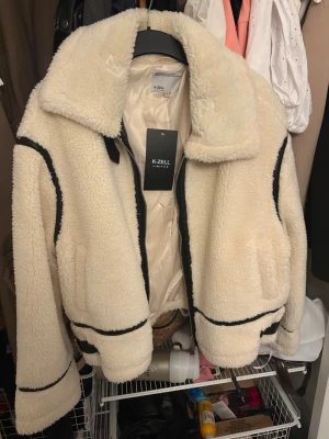 Vit teddyjacka från K-Zell - Snygg vit teddyjacka från K-Zell med svarta detaljer längs dragkedja, ärmar och nederkant. Jackan har stor krage, två fickor och är gjord i mjukt, fluffigt material som ger en riktigt cozy vibe. Perfekt för kyliga dagar när du vill vara både varm och trendig.