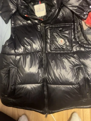 Svart dunväst från Moncler - Snygg svart dunväst från Moncler med glansig finish och klassisk logga på bröstet. Västen har dragkedja framtill, två sidofickor och en bröstficka med lock. Perfekt för lager-på-lager och streetstyle. Materialet är dunfyllning med yttertyg i nylon.