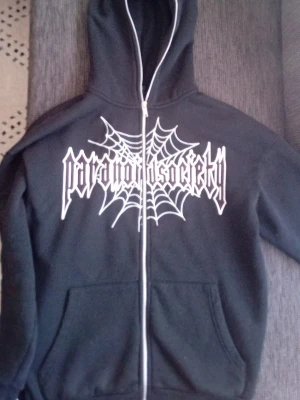 hoodie - Svart hoodie med dragkedja från Paranoid Society. Stor vit tryckt logga och spindelnät på bröstet ger en edgy vibe. Utrustad med huva och två fickor framtill. Perfekt för dig som gillar streetwear och vill sticka ut.