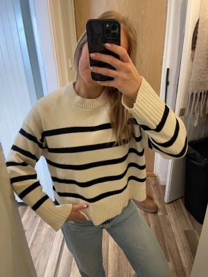 Randig stickad tröja i offwhite och svart - Säljer en stickad tröja i offwhite med breda svarta ränder. Tröjan har en avslappnad passform, rund halsringning och ribbade muddar vid ärmslut och nederkant. Perfekt till jeans för en casual look.