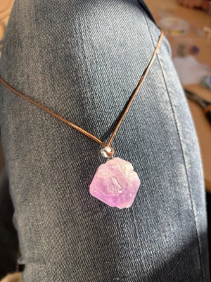 Halsband med lila kristallhänge - Unikt halsband med ett rått, lila kristallhänge på ett brunt läderband. Kristallen har en naturlig, oregelbunden form och ger en bohemisk vibe. Perfekt för dig som gillar statement-smycken och vill sticka ut med en personlig touch.