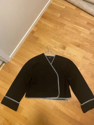 Svart omlottkofta från Gina Tricot - Svart omlottkofta från Gina Tricot med vita dekorativa sömmar längs kanterna. Koftan har lång ärm och en croppad passform som ger en modern look. Perfekt att styla över ett linne eller t-shirt för en trendig outfit. Ena knappen ör av och andra fungerar ej 