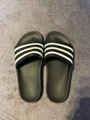 Svarta Adidas badtofflor med vita ränder - Säljer ett par klassiska svarta Adidas tofflor. Har bara använt dem fåtal gånger.