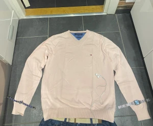 Ljusrosa tröja från Tommy Hilfiger - Snygg ljusrosa tröja från Tommy Hilfiger med V-ringning och diskret logga på bröstet. Tröjan har långa ärmar och ribbade muddar vid ärmslut och nederkant. Perfekt för en stilren och avslappnad look.