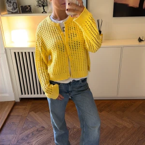 Gul stickad kofta med hålmönster - Trendig gul stickad kofta med hålmönster och vita ribbade kanter. Koftan har knäppning framtill, lång ärm och avslappnad passform. Perfekt för att liva upp din outfit med en färgklick.