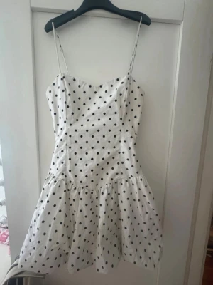 Vit prickig kortklänning med smala band - Supersöt vit kortklänning med svarta prickar och smala axelband. Klänningen har en lätt utställd kjol och figurnära överdel, perfekt för varma dagar. Tillverkad i bomull för en skön och luftig känsla.