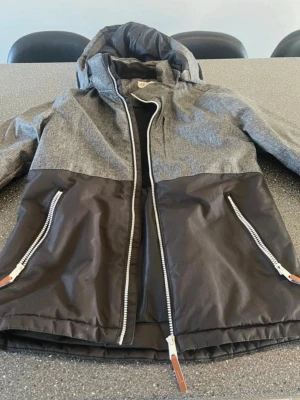 Grå och svart jacka med huva - Snygg tvåfärgad jacka/skidjackq i grått och svart med huva och dragkedja framtill. Jackan har två snedställda fickor med dragkedjor och detaljer. Jackan har en modern, sportig look och funkar perfekt som skidjacka. Storlek 158 13 år. Mycket bra skick.