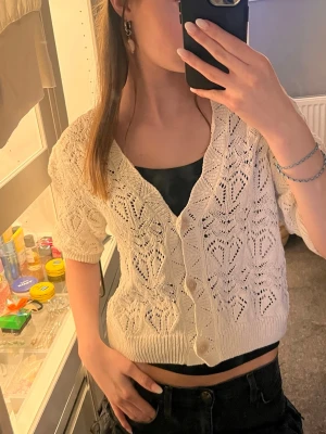 Vit kortärmad stickad kofta - Söt vit stickad kofta med korta ärmar och v-ringning. Koftan har ett fint hålmönster och knäpps med fyra vita knappar framtill. Ribbstickad kant nedtill och vid ärmsluten. Perfekt att slänga över ett linne för en chill look.