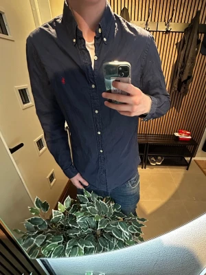 Mörkblå skjorta från Ralph Lauren - Klassisk mörkblå skjorta från Ralph Lauren med knappkrage och vita knappar. Skjortan har lång ärm och den ikoniska röda logotypen broderad på bröstet. Tillverkad i bomull för en bekväm känsla och snygg passform.