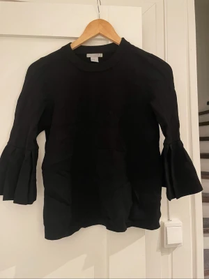 Svart tröja med volangärm från H&M - Snygg svart tröja från H&M i storlek XS. Tröjan har rund halsringning och unika volangärmar som ger en trendig look. Tillverkad i mjukt stickat material som känns bekvämt mot huden. Perfekt för dig som vill ha något stilrent men ändå speciellt.