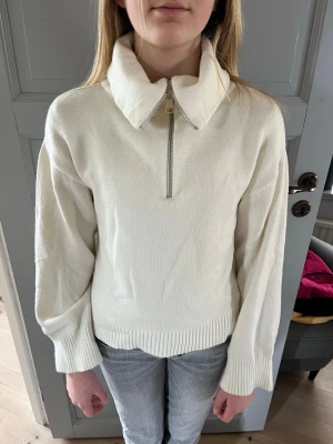 Vit stickad tröja med dragkedja - Säljer en vit stickad tröja med hög krage och dragkedja framtill. Tröjan har långa ärmar, ribbstickade muddar och nederkant. Perfekt för lager på lager och enkel att styla till jeans eller kjol.