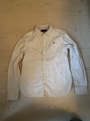 Vit skjorta från Ralph Lauren - Klassisk vit skjorta från Ralph Lauren med button-down krage och broderad logga på bröstet. Skjortan har långa ärmar och knäppning framtill. Perfekt till en stilren och avslappnad look.
