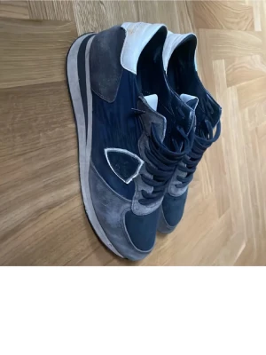 Mörkblå sneakers från Philippe Model - Snygga mörkblå sneakers från Philippe Model med detaljer i grå mocka och vitt läder vid hälen. Skorna har en klassisk låg siluett, vita och grå inslag samt märkets logga på sidan. Perfekta för dig som gillar stilrena och sportiga skor.