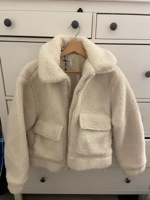 Vit teddyjacka från FB Sister - Säljer en fluffig och supermjuk vit teddyjacka från FB Sister. Jackan har två stora fickor framtill, bred krage och dragkedja. Perfekt för kalla dagar när du vill vara både varm och stylish. Materialet är fuskpäls som ger en mysig känsla.