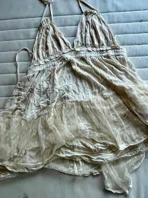 Ljusbeige kort volangklänning - Supersöt kort klänning i ljusbeige med tunna axelband och omlottkänsla. Klänningen har volangdetaljer och ett skirt, transparent lager med diskreta prickar. Perfekt för sommarens varma dagar och ger en romantisk vibe. Materialet känns lätt och luftigt.