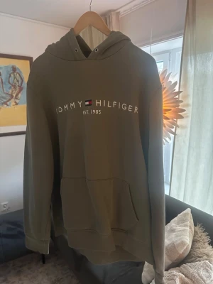Olivgrön hoodie Tommy Hilfiger - Snygg olivgrön hoodie från Tommy Hilfiger med klassisk logga och texten 'EST. 1985' på bröstet. Hoodien har stor känguruficka framtill, justerbar huva med snören och mjukt material som känns skönt mot huden. Perfekt för en avslappnad och trendig look.