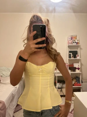 Gul korsettopp med rosett - Söt gul korsettopp med liten vit rosett framtill. Toppen är axelbandslös och har en figurnära passform med markerad midja och lätt utsvängd nederkant. Perfekt för dig som vill ha en trendig och feminin look.