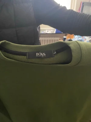 Olivgrön stickad tröja från BOSS - Snygg olivgrön stickad tröja från BOSS med rund halsringning och ribbade muddar vid ärmslut och nederkant. Tröjan har en minimalistisk design och en liten BOSS-logga nertill. Perfekt för dig som gillar stilrena plagg. Aldrig använd.