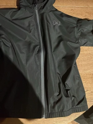 Svart vindjacka från Under Armour - Köpt för 950 kr i wintras aldrig använde prislapp fortfarande på Snygg svart vindjacka från Under Armour med dragkedja framtill och huva. Jackan har en minimalistisk design med logga på bröstet och är tillverkad i ett lätt, vindtätt material som passar perfekt för blåsiga dagar.