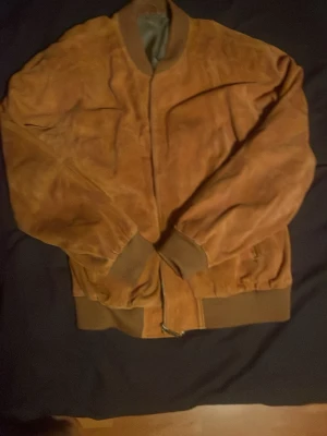 Brun bomberjacka i mocka - Snygg brun bomberjacka i mjuk mocka med ribbade muddar och krage. Jackan har dragkedja framtill och klassisk bomberpassform. Perfekt för dig som vill ha en stilren och trendig look.