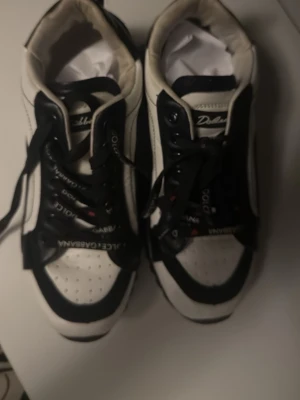 Dolce & Gabbana svartvita sneakers - Snygga svartvita sneakers från Dolce & Gabbana med tydlig logga på hälen och tungan. Skorna har svarta snören med vit text, perforerad tå och kontrasterande detaljer. Tillverkade i läder och textil för en exklusiv känsla och modern look.