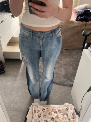 Blå bootcut jeans från Lee - Säljer ett par blå bootcut jeans från Lee med klassisk femficksmodell och ljus tvätt. Jeansen har en patchad detalj på vänster lår och Lee-logga på bakfickan. Tillverkade i bomull med en skön passform och normal midja. Perfekta till en avslappnad stil.
