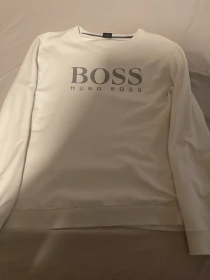 Vit sweatshirt från Hugo Boss - Snygg vit sweatshirt från Hugo Boss med stor logga på bröstet. Tröjan har rund halsringning och långa ärmar. Perfekt för en clean och stilren look. Tillverkad i mjukt material för extra komfort. Har ett litet hål som man kan se på armen där men ej så synligt