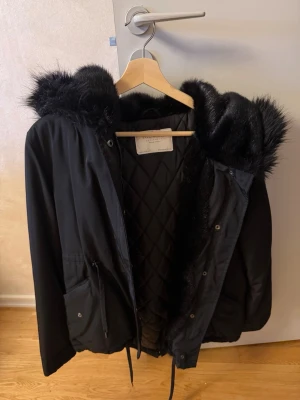 Svart parkas med fuskpäls från Zara - Snygg svart parkas från Zara med stor huva kantad av mjuk svart fuskpäls. Jackan har quiltat innerfoder, dragsko i midjan och praktiska fickor framtill. Perfekt för kalla dagar när du vill vara både varm och stilren.