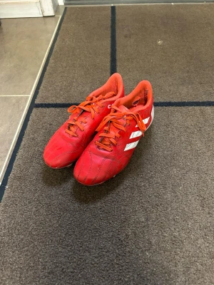 Röda Adidas fotbollsskor med vita ränder - Säljer ett par röda fotbollsskor från Adidas med klassiska vita ränder på sidan. Skorna har snörning och låg profil, tillverkade i syntetmaterial för bra bollkontroll. Perfekta för dig som vill sticka ut på planen!
