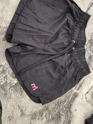 Svarta träningsshorts från Mistral - Svarta träningsshorts från Mistral med elastisk midja och snörning. Tillverkade i 100% polyester och har en liten rosa logga på benet. Perfekta för sportiga aktiviteter och har en loose passform för extra rörelsefrihet.