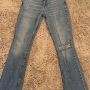 Bootcut jeans från Gina tricot, ljusblå - Ljusblå bootcut jeans från Perfect Jeans, Gina tricot med slitning och hål på ena benet. Klassisk femficksmodell, låg midja och råa kanter nertill. Jeansen är tillverkade i bomull. Säljer pågrund av att de är för långa så dem kommer inte riktigt till användning.