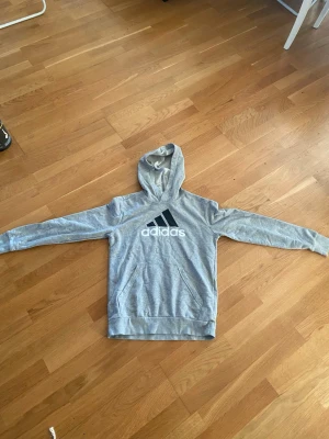 Grå adidas hoddie  - Säljer en grå hoodie från Adidas med stor svart logga och vit text på bröstet. Hoodien har huva, magficka och ribbade muddar vid ärmar och nederkant. Tillverkad i mjukt sweatshirtmaterial, perfekt för en avslappnad stil.